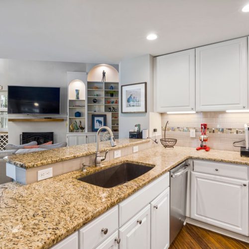 A modern kitchen with granite countertops opens into a stylish living room, featuring elegant furniture and built-in shelves.