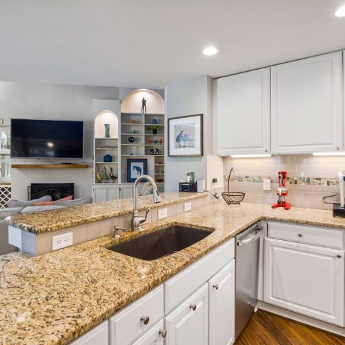 A modern kitchen with granite countertops opens into a stylish living room, featuring elegant furniture and built-in shelves.